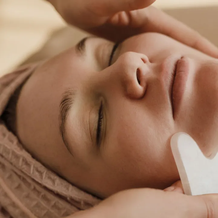 Terapis memijat wajah pelanggan dengan batu gua sha; pelanggan berbaring dengan mata terpejam dan rambut dibungkus handuk