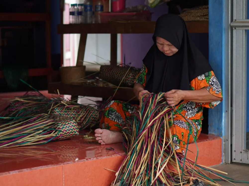 A woman in a black headscarf sits on her porch step, weaving a bundle of long purun strips in green, pink and natural