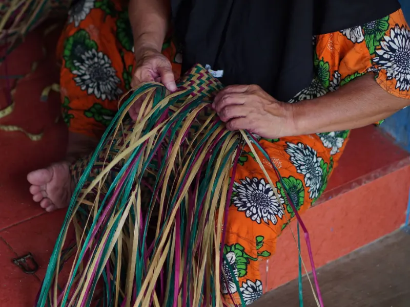 Hands sorting long strips of green, pink and natural purun on a red-tiled porch