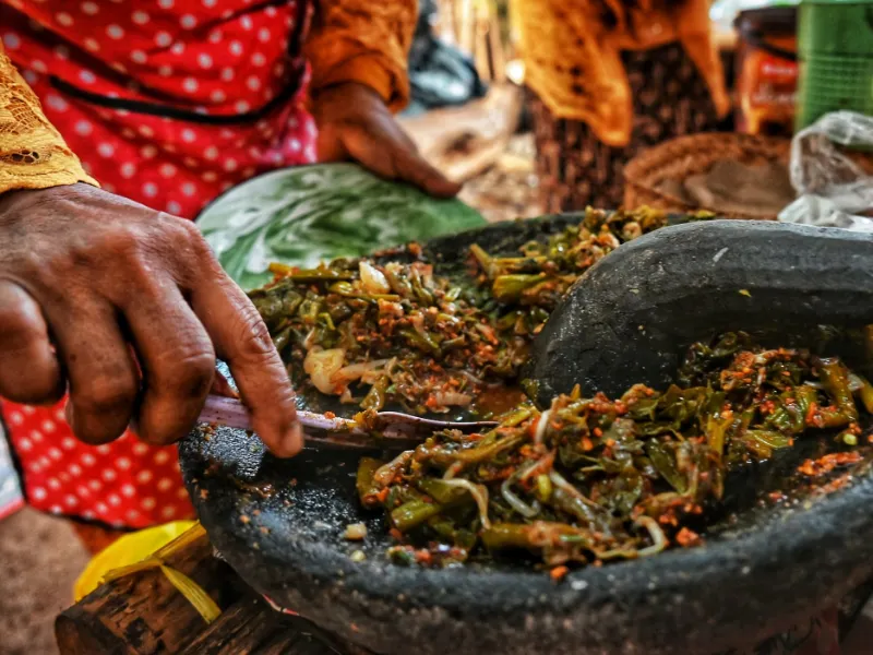 Hands grinding chilli and leaves with a stone pestle in a wide stone mortar